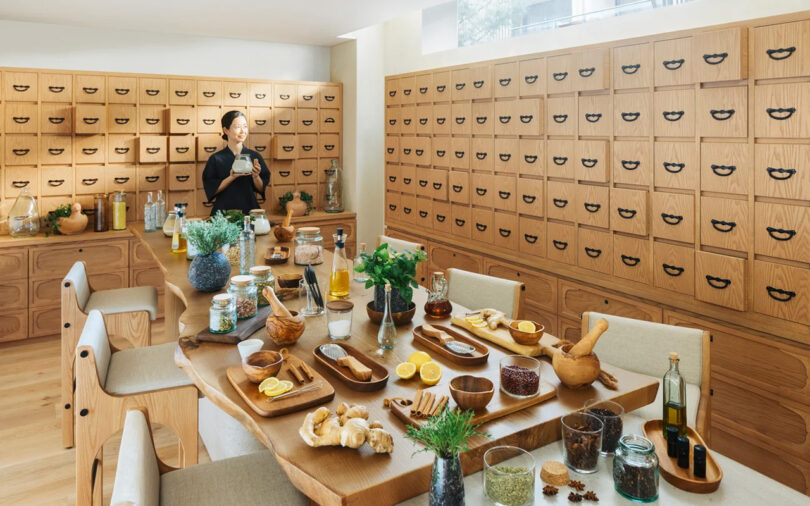 A woman preparing herbs in a modern kitchen at the Six Senses Kyoto hotel, with numerous wooden drawers and a dining table set with various ingredients and dishes.