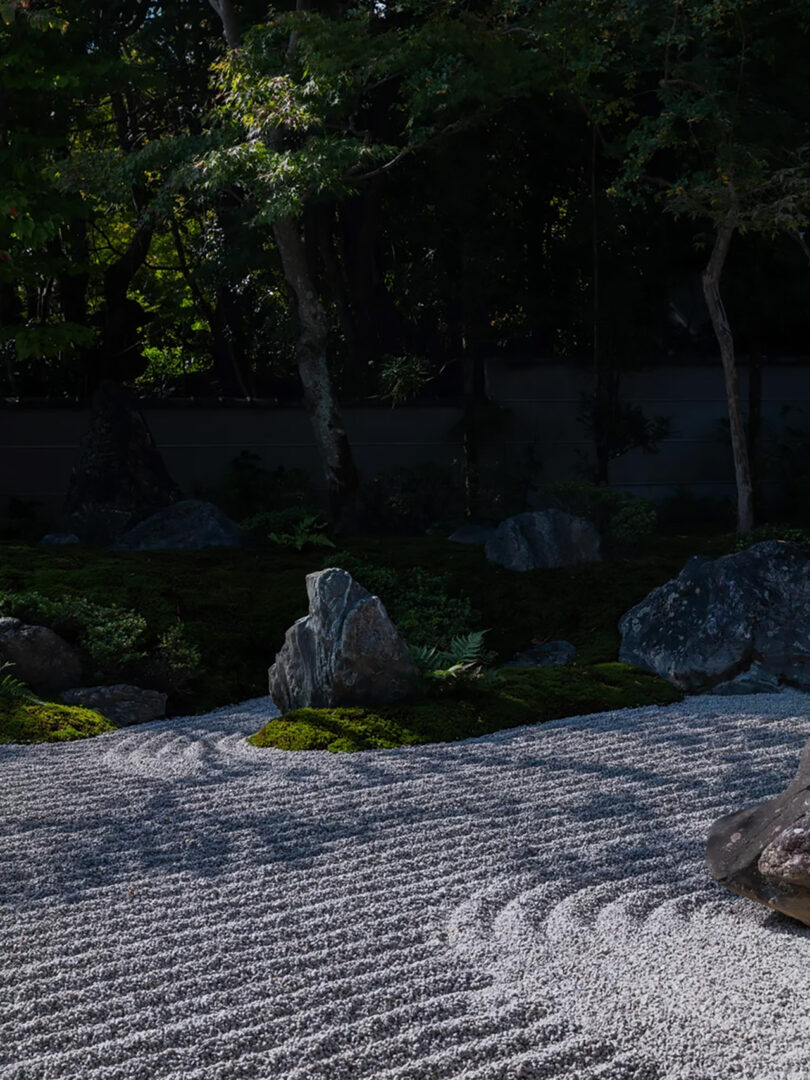 A serene Japanese rock garden at the Six Senses Kyoto hotel with meticulously raked gravel, moss, and strategically placed rocks, surrounded by shady trees.