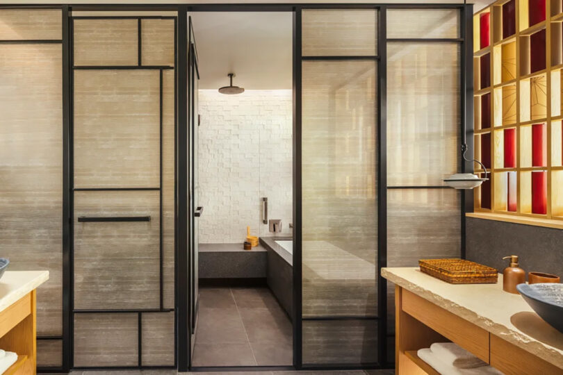 Modern bathroom at Six Senses Kyoto hotel with sliding doors, featuring a bathtub, white tiled walls, and a wooden shelving unit.