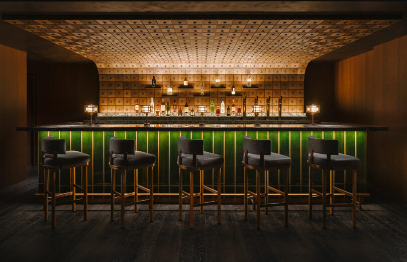Dark moody illuminated bar with green paneled front, curved ceiling, and five black and gold legged bar stools angled toward the right side of the room.