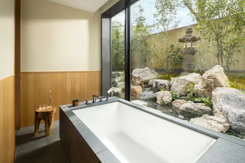 Modern bathroom with large bathtub, glass wall overlooking a serene Japanese garden at the Six Senses Kyoto hotel, featuring a stone fountain and lush greenery.