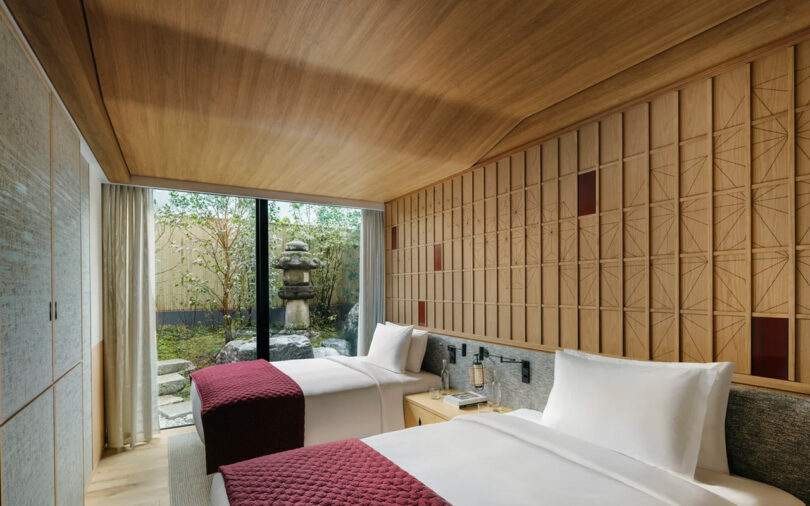 Modern bedroom at the Six Senses Kyoto hotel featuring wooden panels, a patterned wall, two beds with red accents, and a tranquil Japanese garden view through large windows.