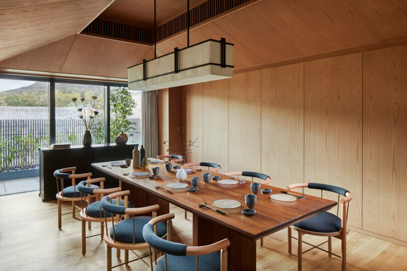 Modern dining room at the Six Senses Kyoto hotel with a wooden table set for six, blue chairs, large hanging lamp, and panoramic windows overlooking a balcony with trees.