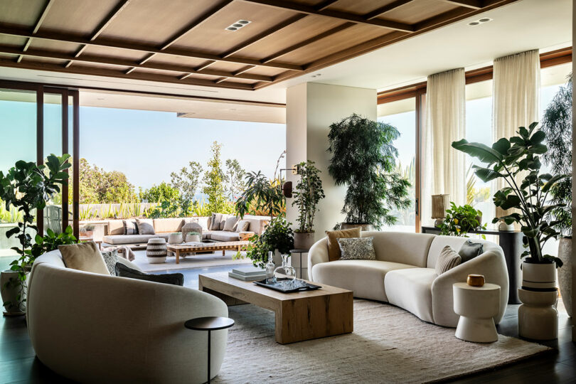 Luxurious living room designed by SkB Architects with modern decor, large windows, and potted plants, opening onto an outdoor patio area.