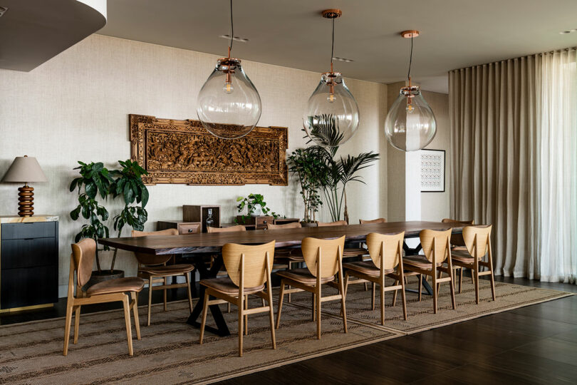 Modern dining room designed by SkB Architects featuring a long wooden table with chairs, ornate wall art, large pendant lights, and indoor plants.