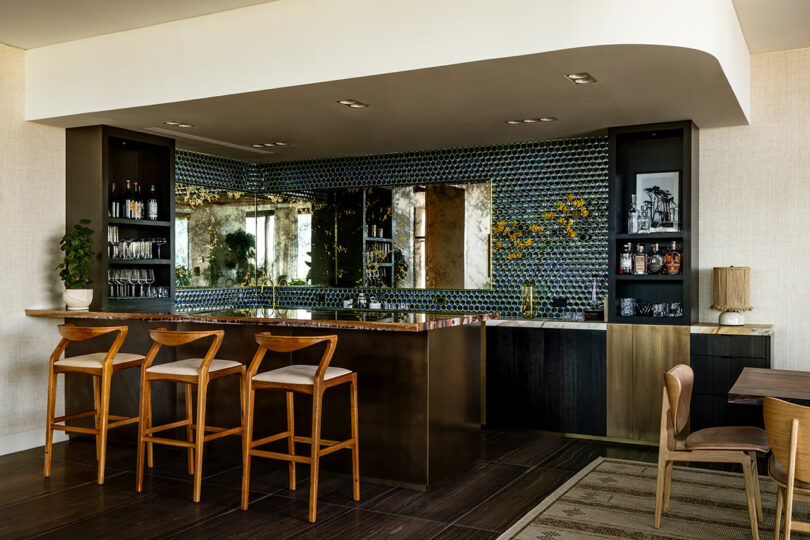 Elegant bar area designed by SkB Architects with a mirrored backsplash, patterned tiles, wooden stools, and shelves displaying bottles and glasses.