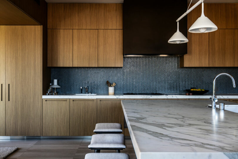 Modern kitchen interior designed by SkB Architects, featuring wooden cabinets, white marble countertops, and stylish pendant lights over a breakfast bar with stools.