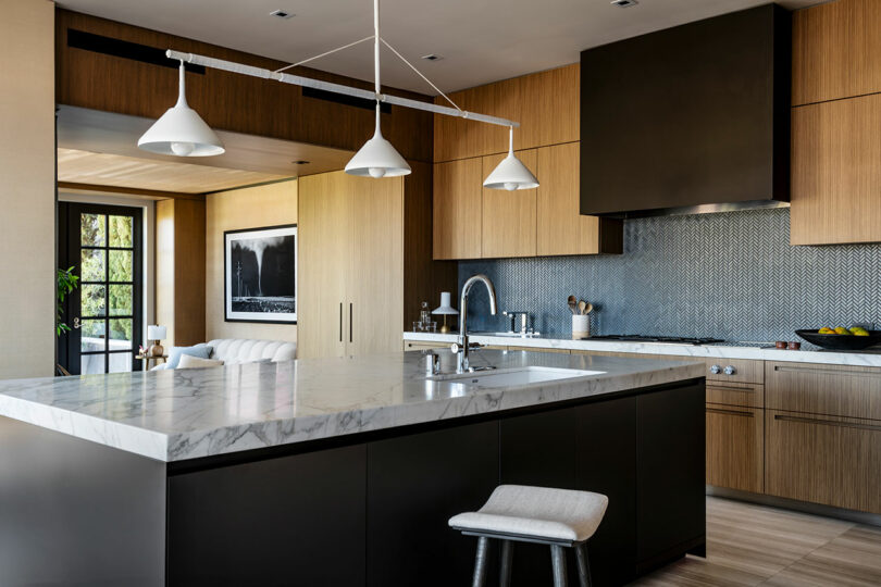 Modern kitchen interior designed by SkB Architects with wooden cabinets, a large black island with marble countertop, pendant lights, and a view to the outdoors through glass doors.