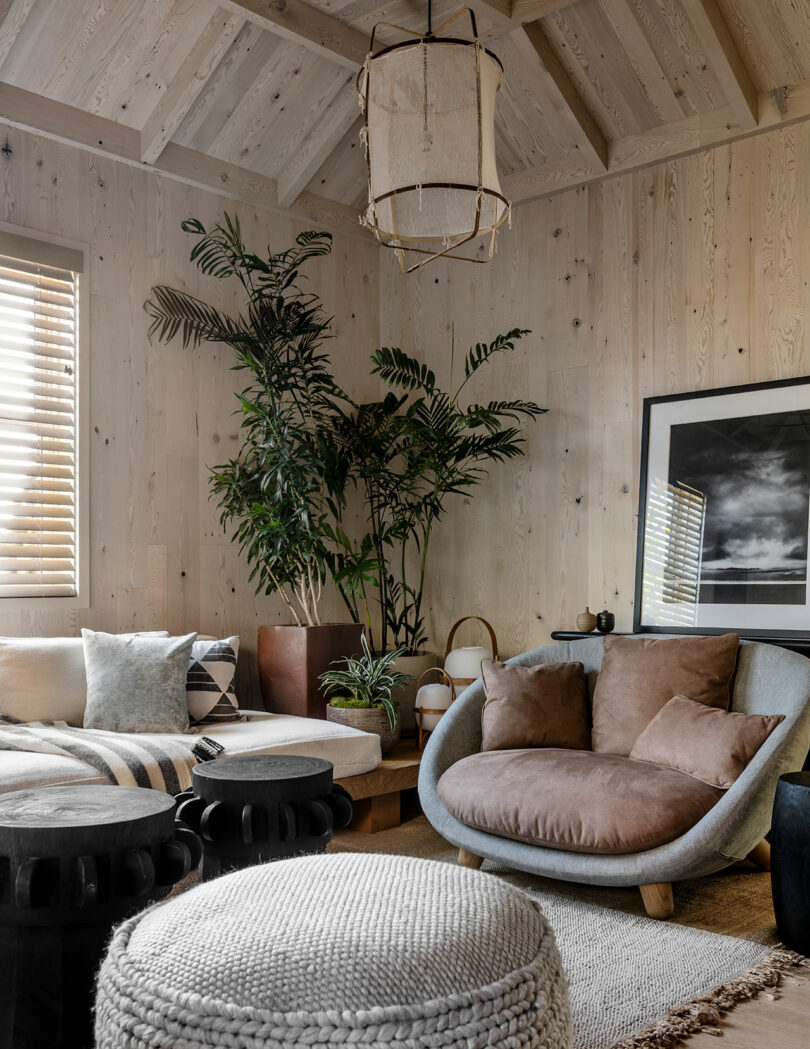 A cozy living room designed by SkB Architects with a beige sofa, round chairs, wooden walls, an assortment of plants, and a hanging light fixture.