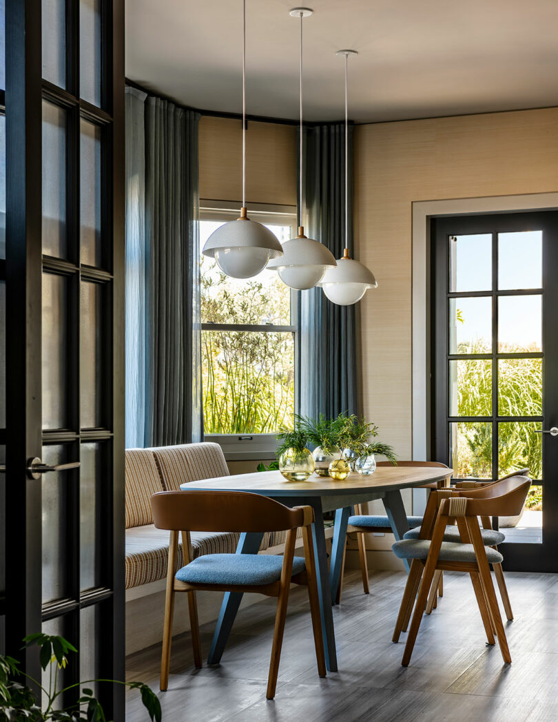 Modern dining room designed by SkB Architects with a wooden table, blue and beige chairs, and three pendant lights, overlooking a garden through large windows.