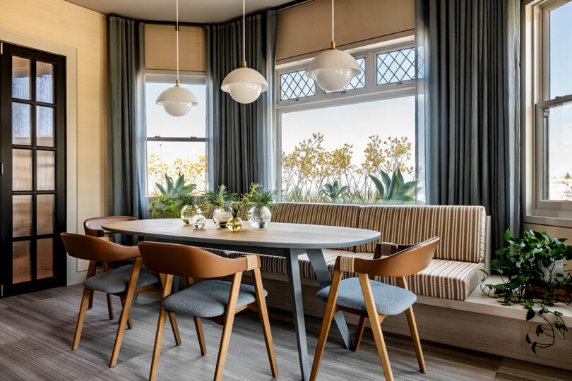 Modern dining room designed by SkB Architects, featuring a curved booth, wooden table, blue chairs, pendant lights, and large windows with yellow flowers on the sill.