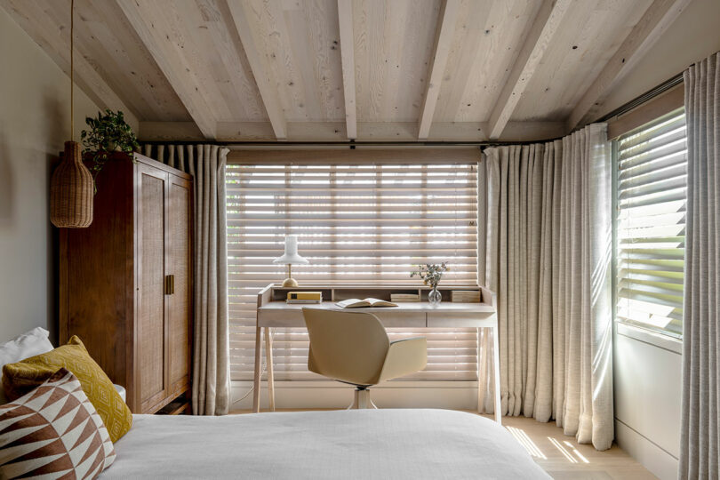 A modern bedroom designed by SkB Architects with a wooden ceiling, large windows with blinds, a desk with a chair, and a plant hanging in a basket.