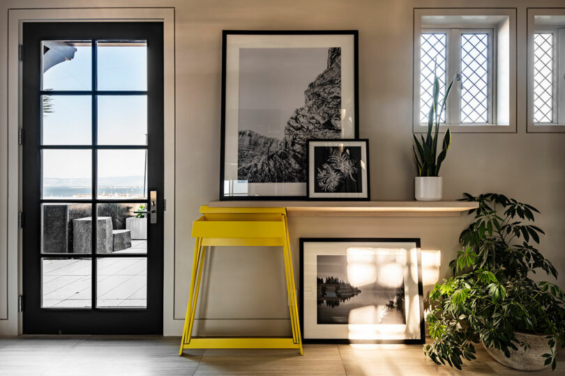 Modern entryway designed by SkB Architects featuring a black door, a yellow stool, black console table, framed pictures, and plants beside a sunny window.