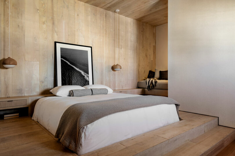 Modern bedroom designed by SkB Architects, featuring a minimalist aesthetic with a large bed adorned in gray bedding, wooden walls, and a framed black-and-white beach photograph.