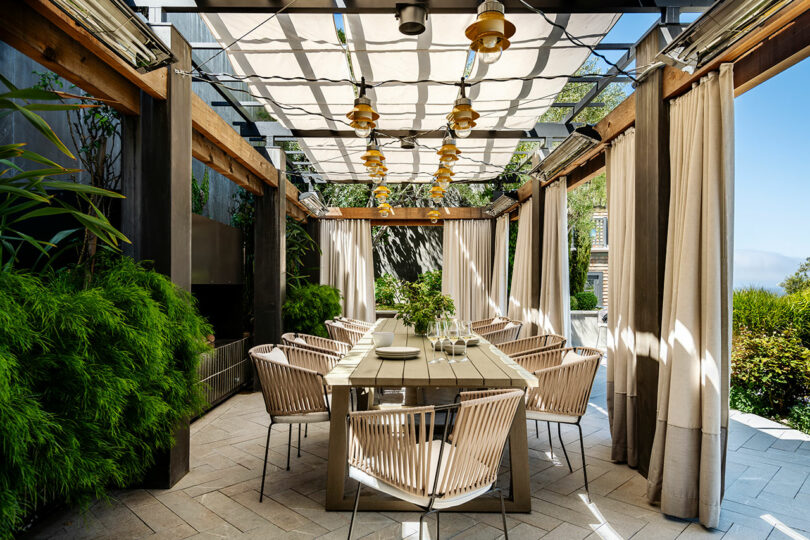 Outdoor dining area designed by SkB Architects with wooden tables, chairs under a pergola with hanging lights and sheer curtains, surrounded by lush greenery.
