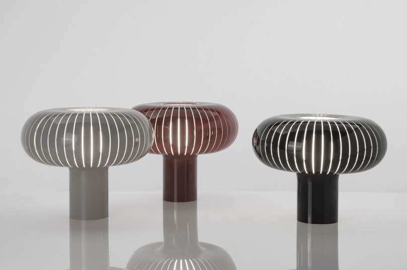 Three modern, ribbed table lamps in gray, maroon, and black colors side by side on a reflective surface against a white background.