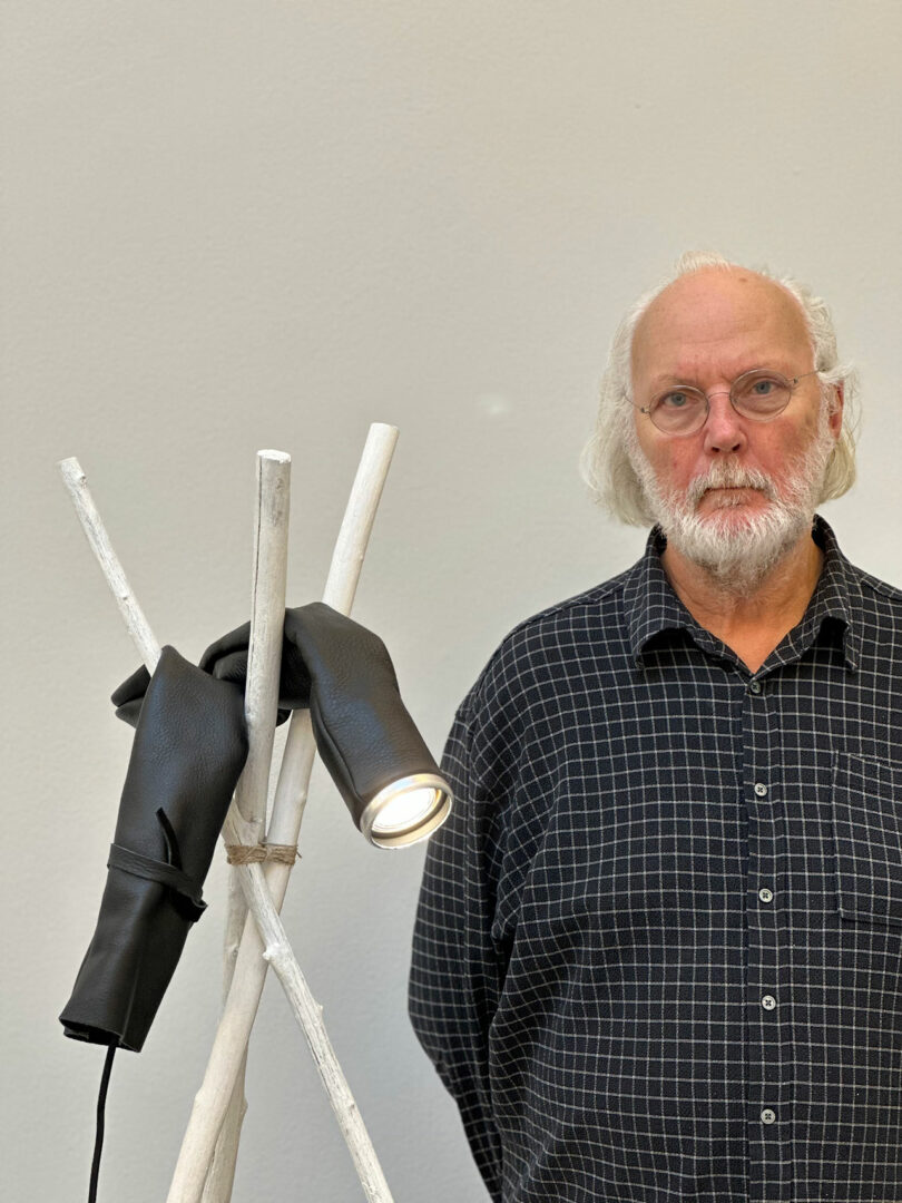 An older man with a beard standing next to an upright tripod with a sinuous flashlight attached to it.