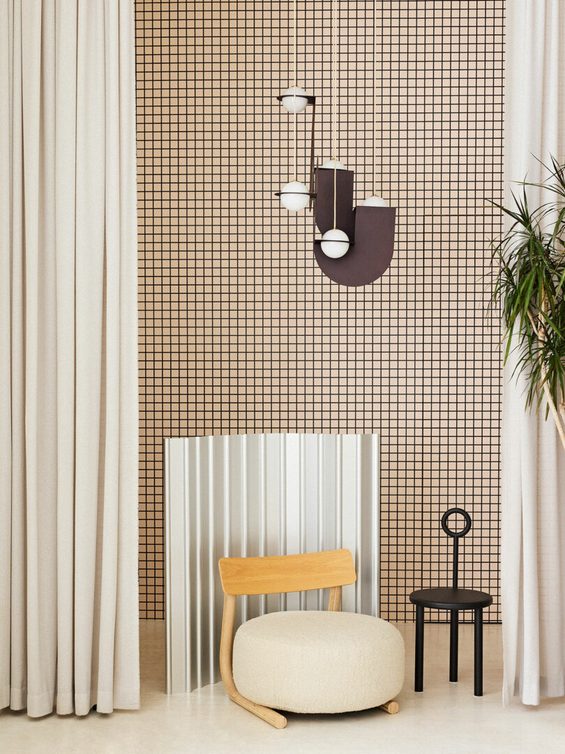 A modern living room corner featuring a beige armchair, a small black side table, a corrugated white panel, and a geometric-patterned wall with a contemporary wall lamp.