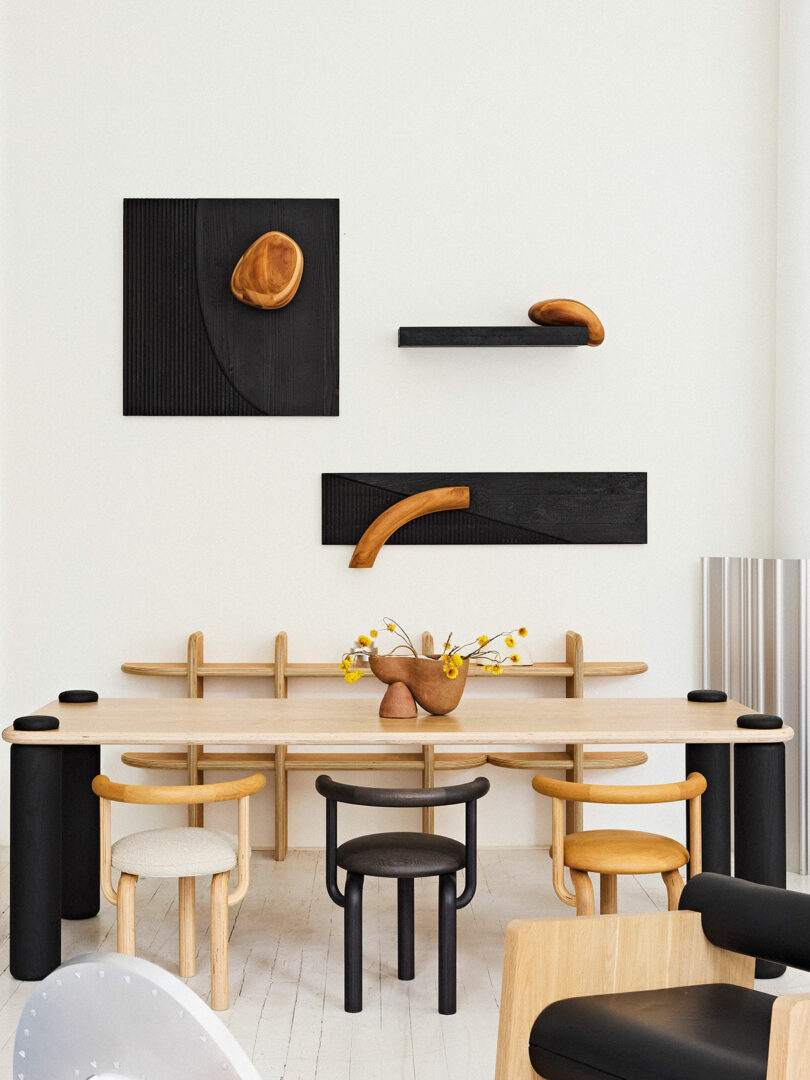 Minimalist dining room with a wooden table, mixed chairs, and abstract black wall art featuring curved forms. shelves adorned with unique sculptures and a small vase of flowers add charm.