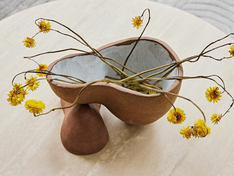 A ceramic bowl with a unique curved design holds dried yellow flowers on a wooden table.