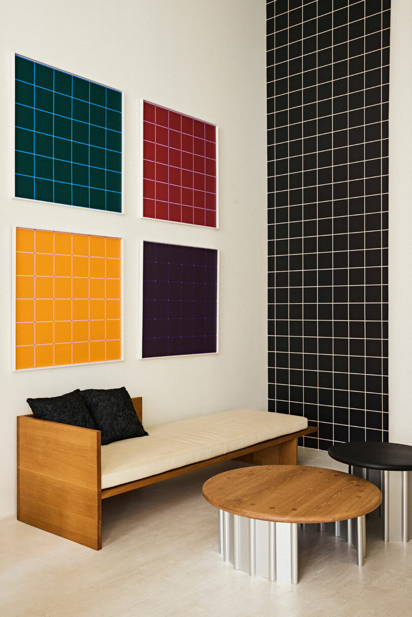 Modern interior featuring a wooden bench with black cushions, a wooden circular table, and a black table against a white wall adorned with colorful grid-patterned panels.