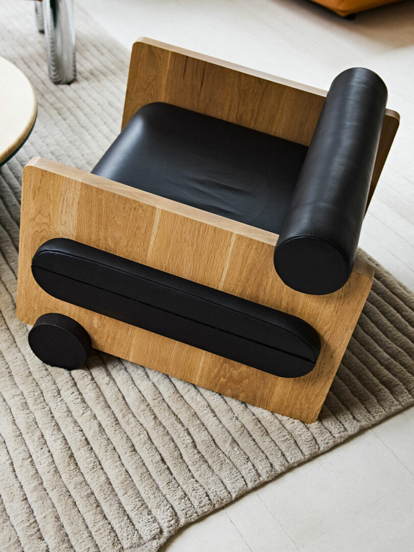 A modern wooden armchair with black leather cushions and cylindrical elements, positioned on a beige rug in a room with light flooring.