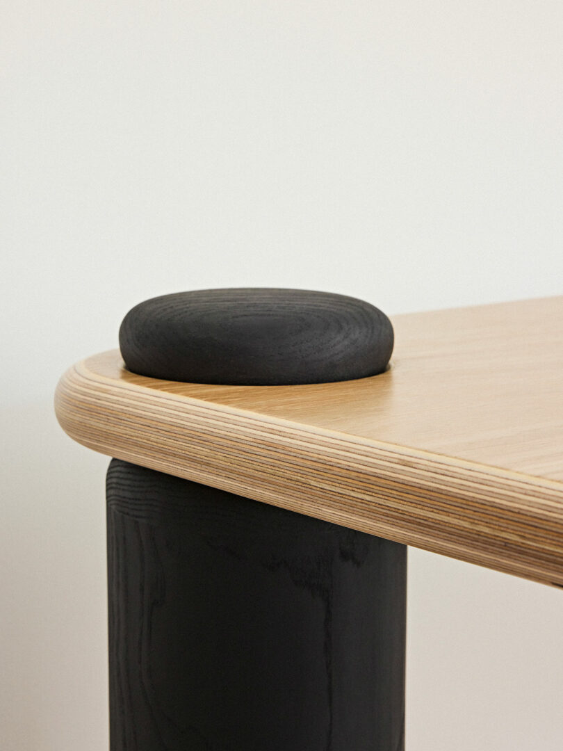 Close-up of a modern table corner with a rounded black cushion on top of a black cylindrical leg and a light plywood tabletop.