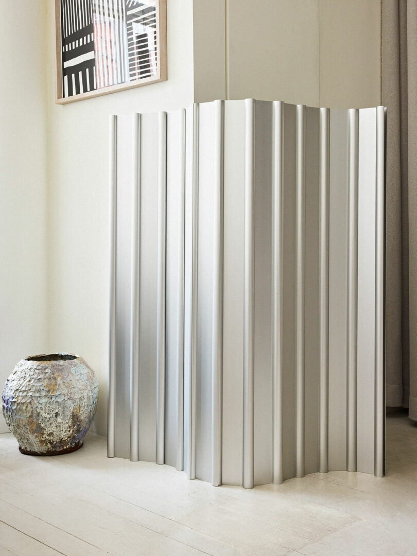 A tall, white, vertical pleated room divider next to a large, textured vase in a bright, minimalistic room.