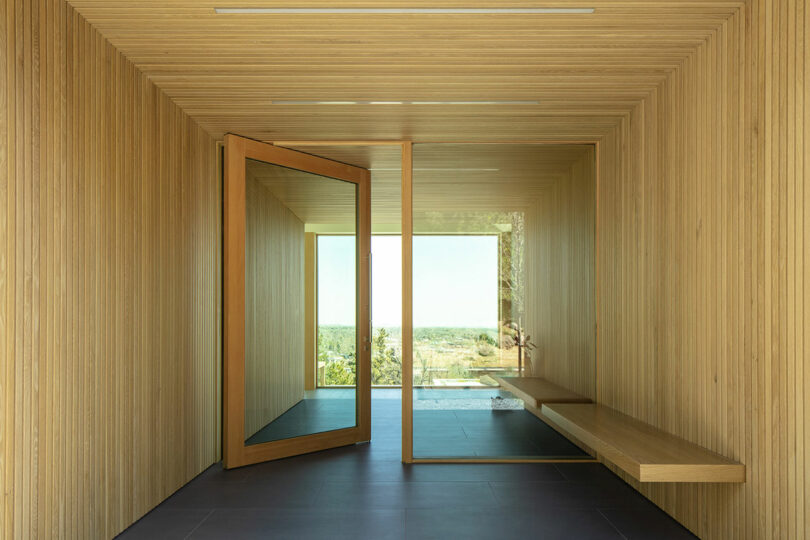 A minimalist wooden hallway in Villa H with a large glass door open to a natural landscape, featuring a bench on the right and a glass ceiling strip allowing natural light.