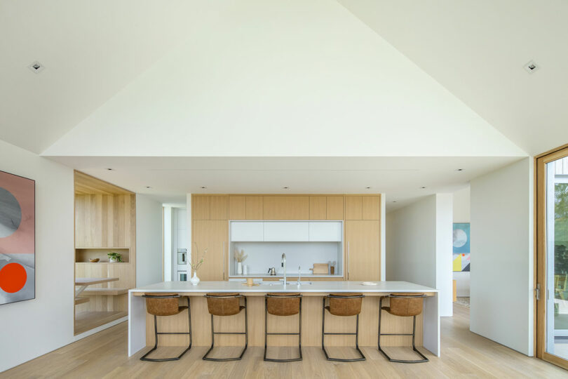 Modern kitchen in Villa H with a minimalist design featuring wooden cabinets, a central island with bar stools, large windows, and a geometric ceiling.