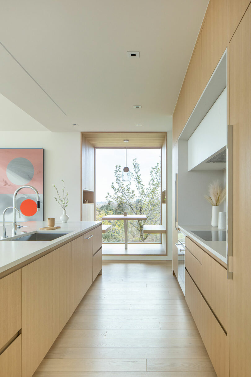 Modern kitchen in Villa H with light wood cabinets, a clean countertop with minimal decor, and a large window providing natural light and a view of trees.