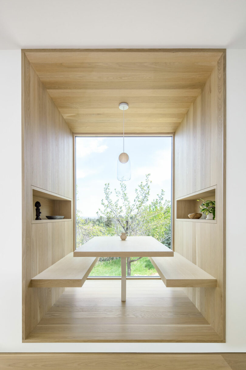 A minimalist reading nook in Villa H with wooden built-in benches and shelves, overlooking a garden through a large window, lit by a simple pendant light.