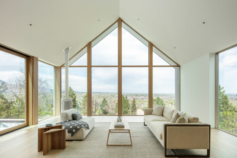 Modern living room in Villa H with large triangular windows offering a panoramic view of a mountainous landscape, furnished with neutral-toned sofas and minimalist decor.