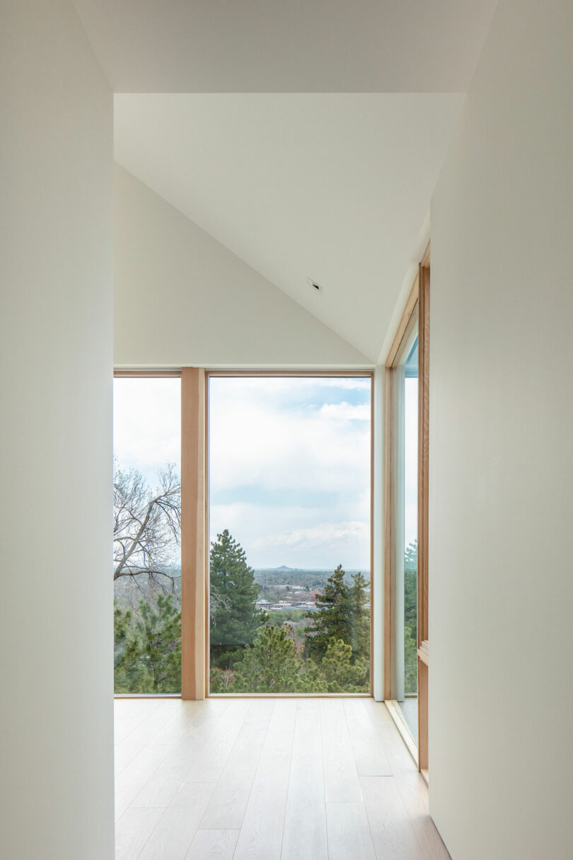 Interior view of Villa H, featuring a minimalistic hallway with wooden floors leading to a large window showcasing a scenic view of the countryside.