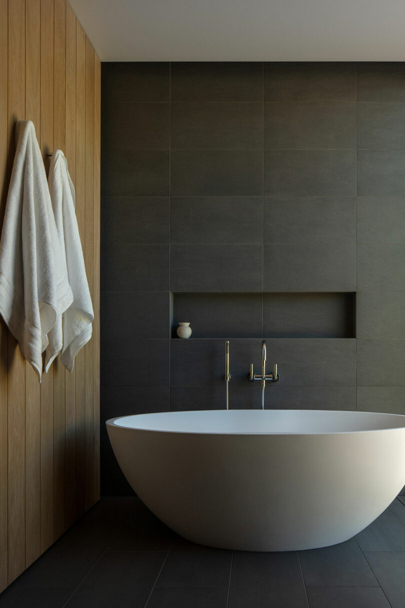 Modern bathroom in Villa H features a freestanding oval bathtub, wooden wall paneling, and gray tiled walls. Towels hang on the side with minimalist fixtures.