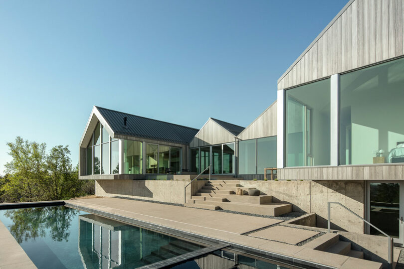 Villa H with large glass windows, asymmetric roofs, and an adjacent swimming pool, surrounded by a natural landscape under a clear sky.