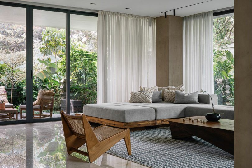 Modern living room with a large gray sectional sofa, wooden chairs, floor-to-ceiling windows, sheer curtains, and lush greenery outside.