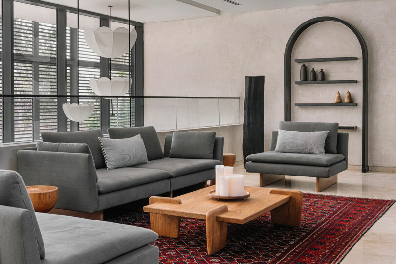 Modern living room with gray sofas, wooden tables, an arch-shaped niche, and pendant lights, featuring large windows and a textured wall.