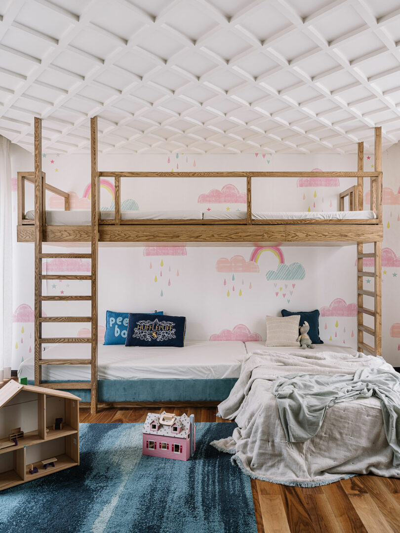 A cozy children's bedroom with a wooden bunk bed, a cloud and raindrop themed wallpaper, and a small play area on a blue rug.