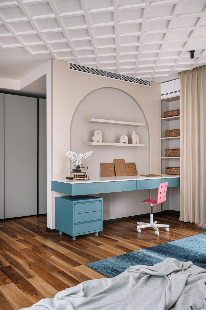 A modern home office with a light blue desk, pink chair, and built-in shelves against a textured white wall, with a wooden floor and a blue rug.
