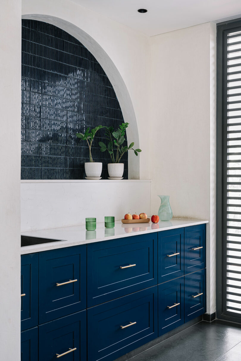 A modern kitchen corner featuring blue cabinets, a white countertop, and a large arched window with a grid pattern. potted plants and fruit are neatly arranged on the surfaces.