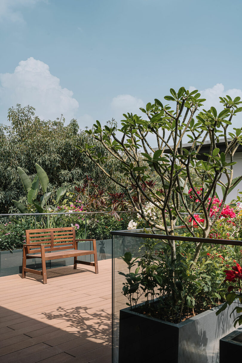 A serene rooftop garden with a wooden bench, lush green plants, and vibrant flowers, surrounded by clear glass balustrades under a clear sky.