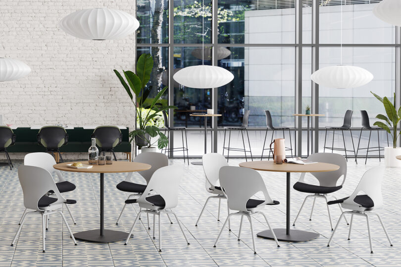 Modern office breakroom featuring round tables with white and gray chairs, large windows, and pendant lights in a spacious, plant-decorated space.