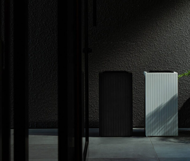 Two rectangular trash bins, one gray and one white, stand against a textured dark wall between shadowed square pillars.