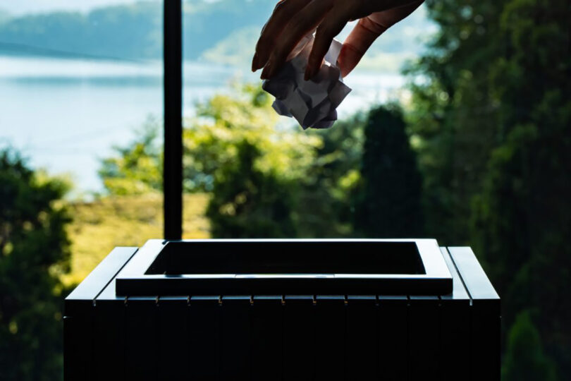 A hand dropping a crumpled paper into a trash can, with a scenic lake and greenery visible in the background through a square window.