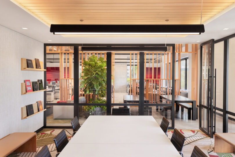 Modern office meeting area with a long white table surrounded by patterned chairs, wooden slatted partitions, ample natural light, and lush indoor plants