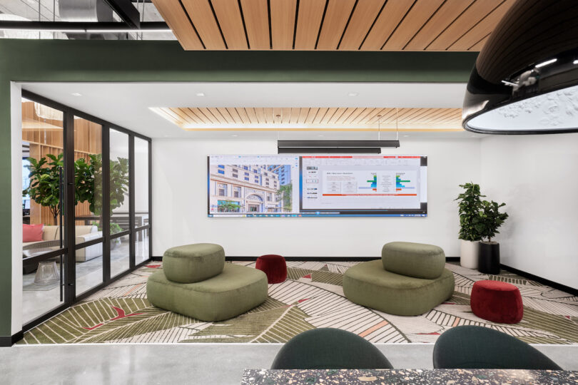 A modern office lounge space with green seating, a large digital display screen, wooden ceiling panels, and indoor plants.