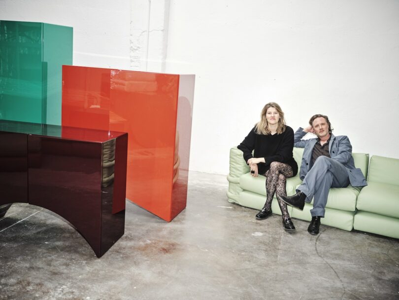 a man and woman sitting on a mint green sofa next to three colorful cabinets