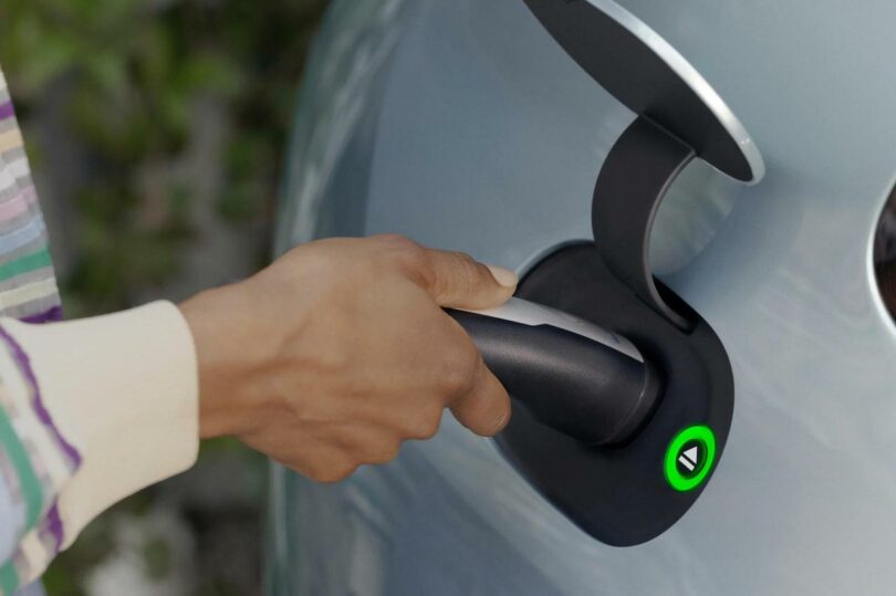 A person's hand plugging a charger into an electric vehicle's charging port, indicated by a glowing green light