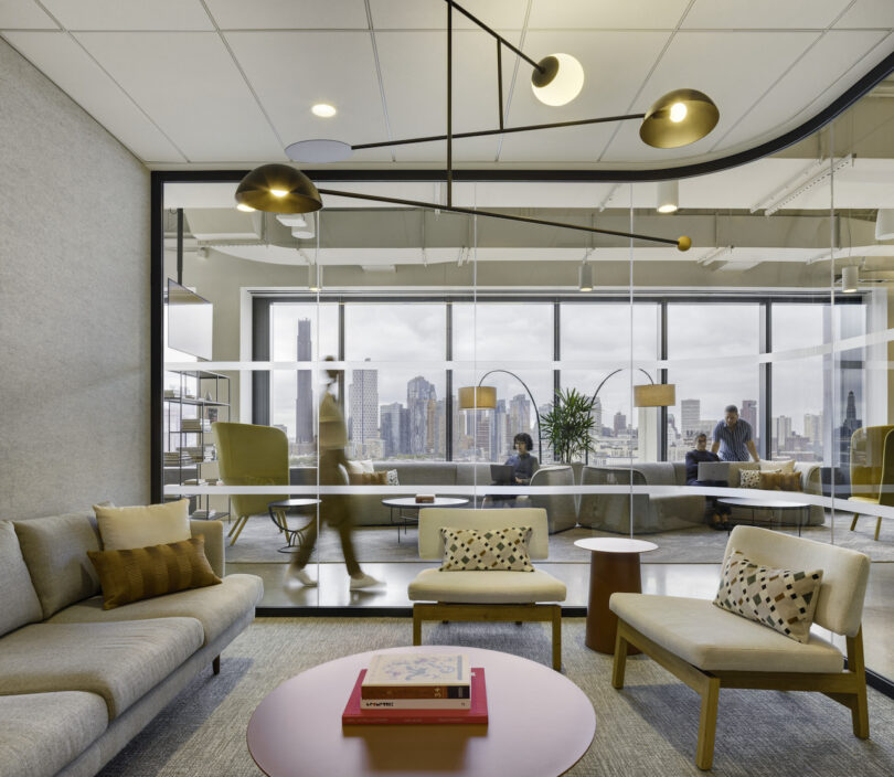 Modern office lounge with comfortable seating, stylish lighting, and a city view, occupied by several people working and conversing.